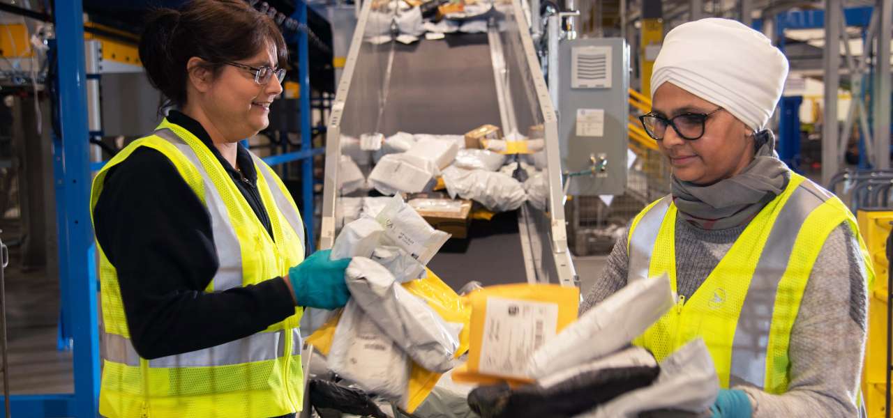 Two female Canada Post employees wearing bright yellow safety vests sort through packages and envelopes.