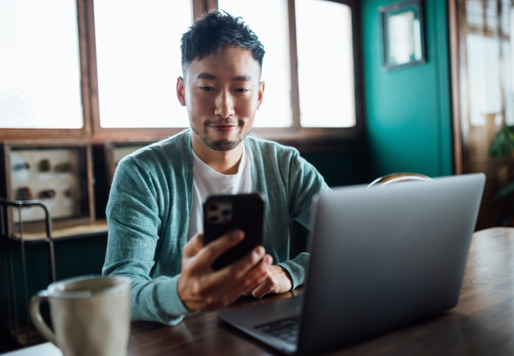 A man looks at his cellphone while sitting in front of a laptop at a wooden table.