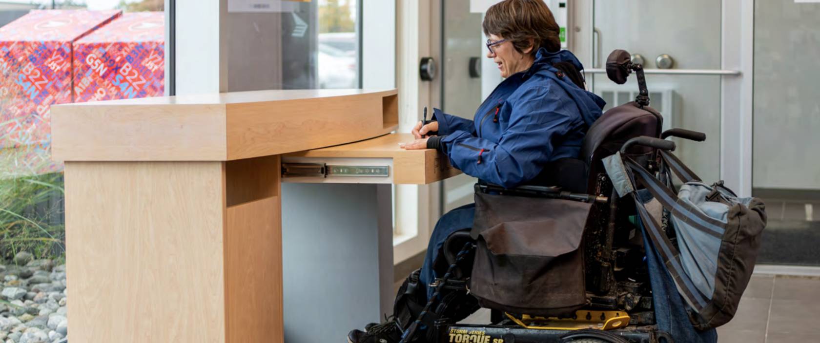 Une femme en fauteuil roulant remplit un formulaire à un bureau de poste accessible.