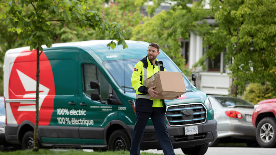 Un conducteur de Postes Canada transporte un colis, sa fourgonnette 100 % électrique stationnée sur une rue derrière lui. 